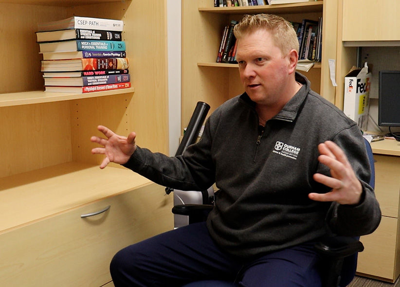 A man with blonde hair and a grey quarter-zip sweater sits in a desk chair and gestures with his hands in the middle of the image. To his left is a beige shelf with health textbooks and behind him a desk and additional shelves full of books and a computer.