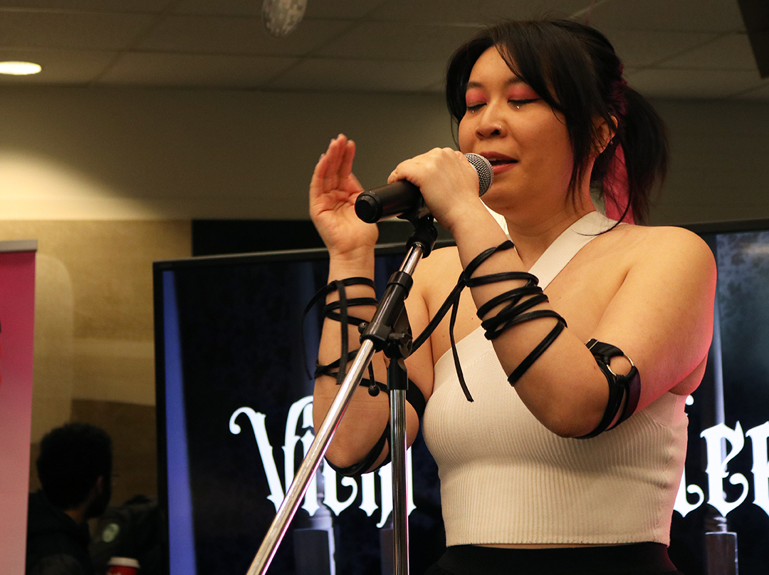 A woman with long dark hair pulled into a ponytail sings into a microphone. She is wearing a white sleeveless shirt.