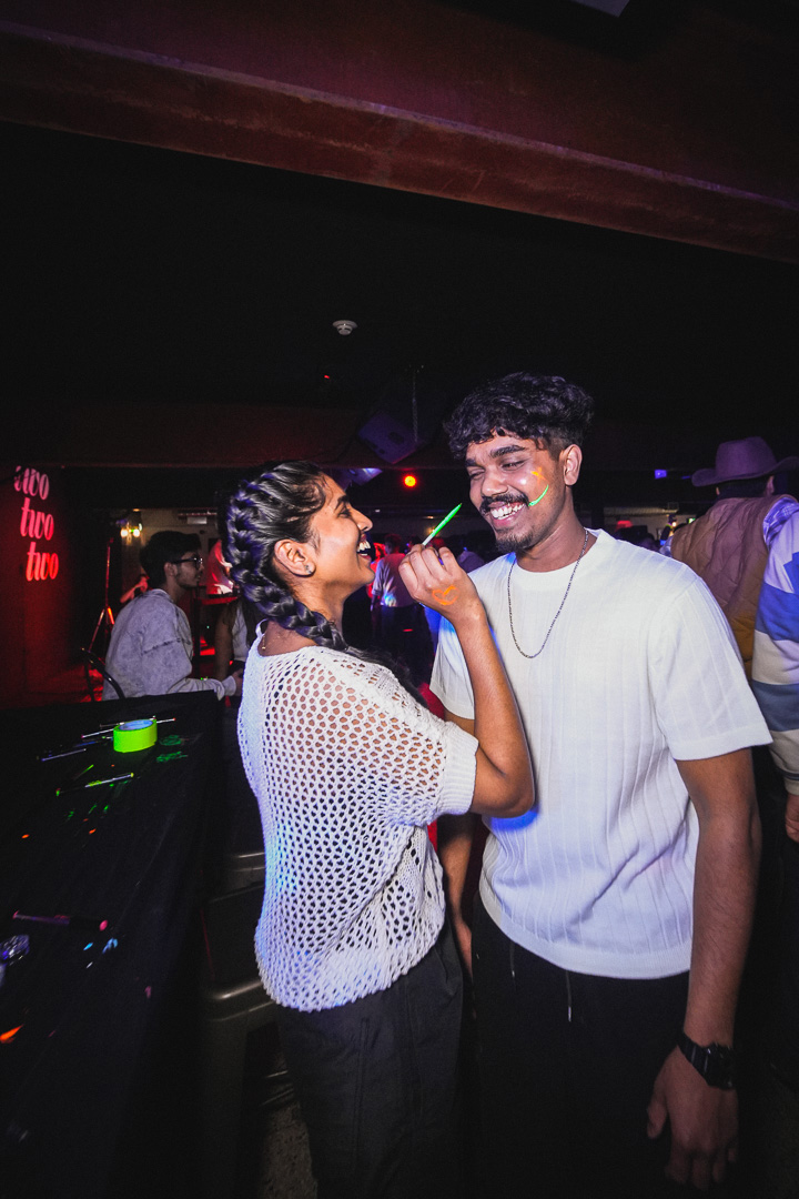 A young woman in a white shirt paints the face of a man in a white shirt. The woman is holding a green paintbrush.