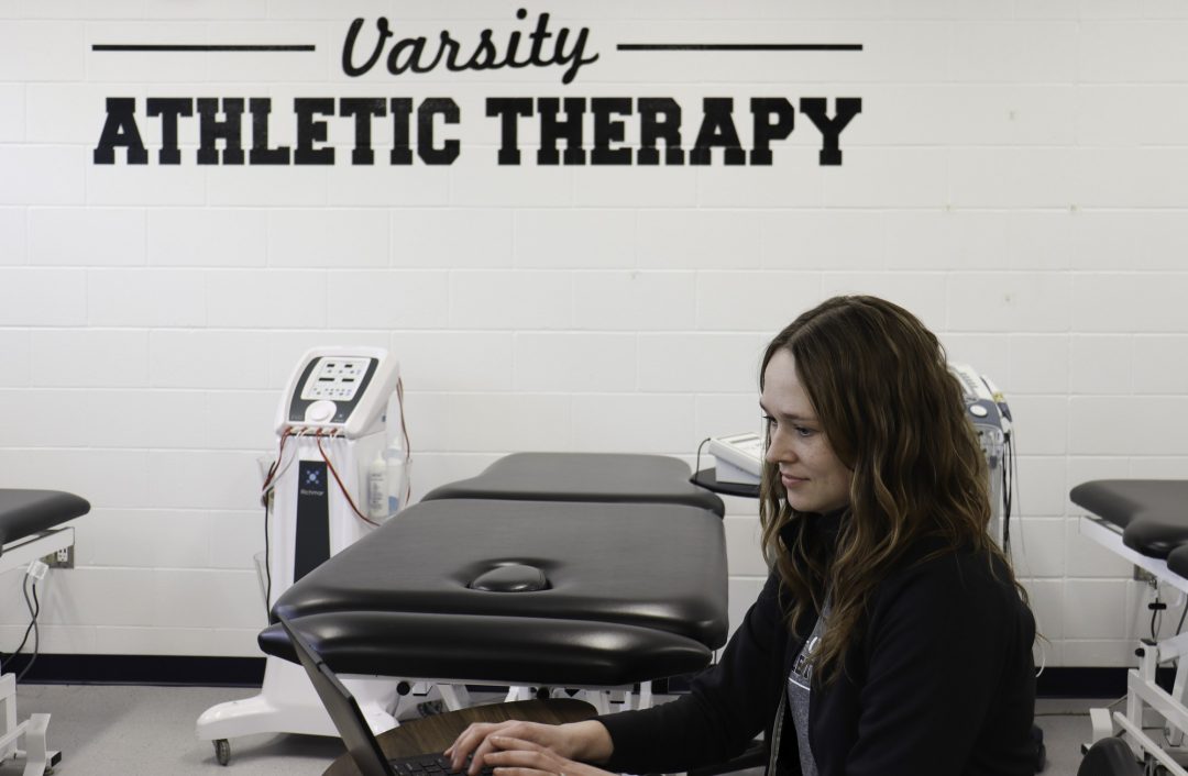 A woman is sitting at a table typing on a laptop inside of Durham College's athletic therapy clinic.