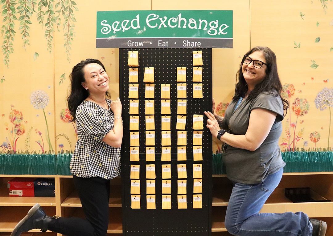 Two women stand between a tall board holding a variety of seed pouches. At the top is a sign that says "seed exchange."