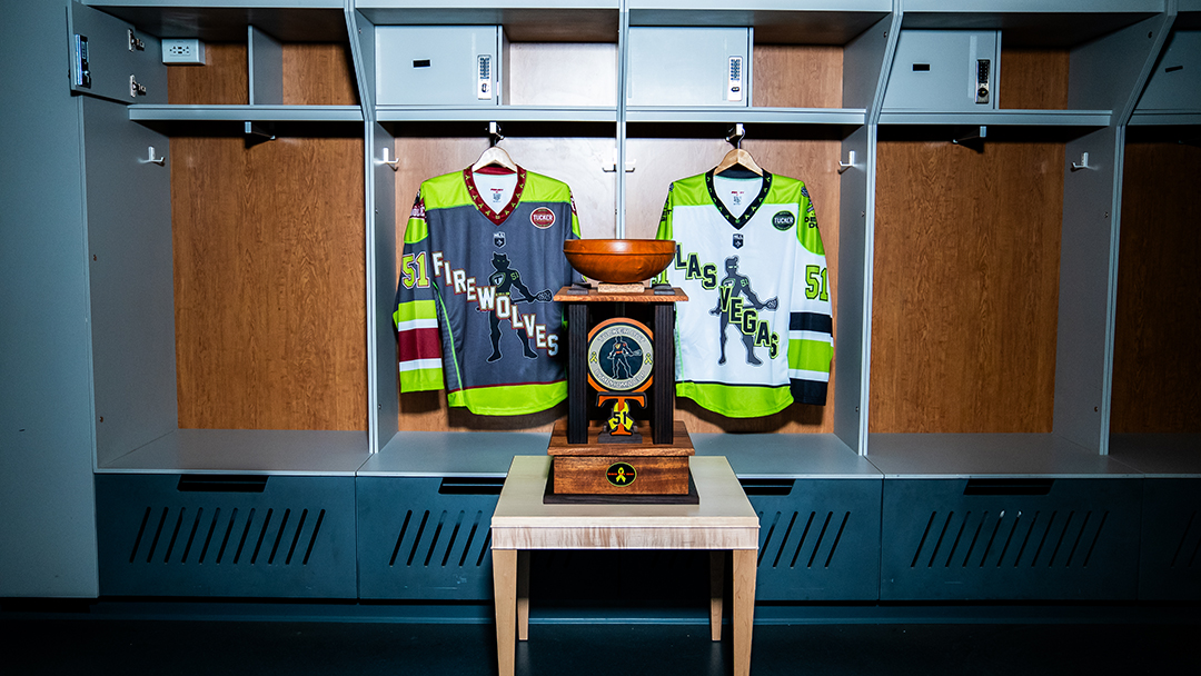 A trophy sitting on a table in a locker room in front of two jerseys, each from a different team.