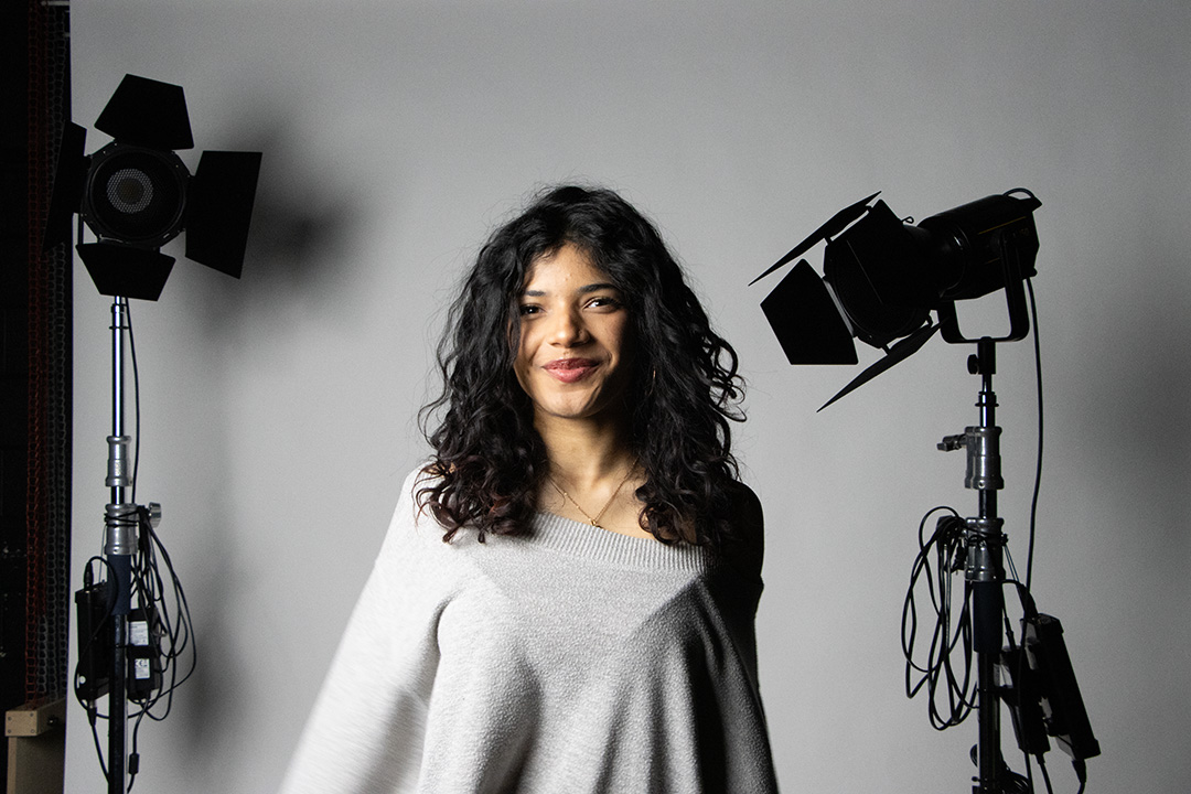 A young woman poses for a photo. She has black curly hair and a grey off-shoulder sweater, with a grey background and photography equipment on either side of her.