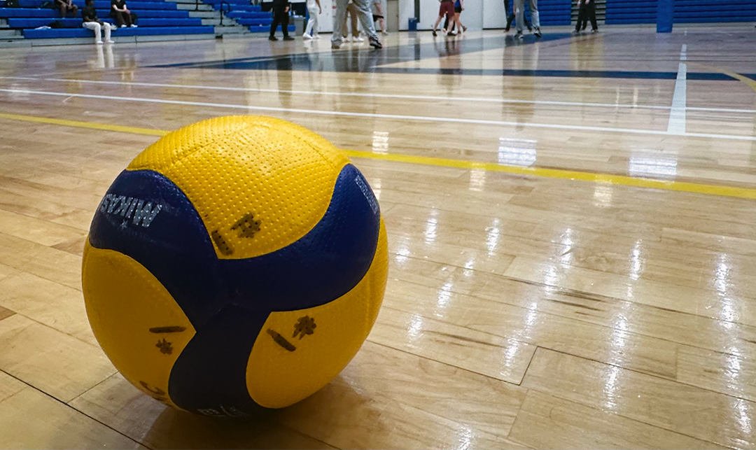 A yellow and blue volleyball sits on a basketball court with feet of students playing the game in the background.