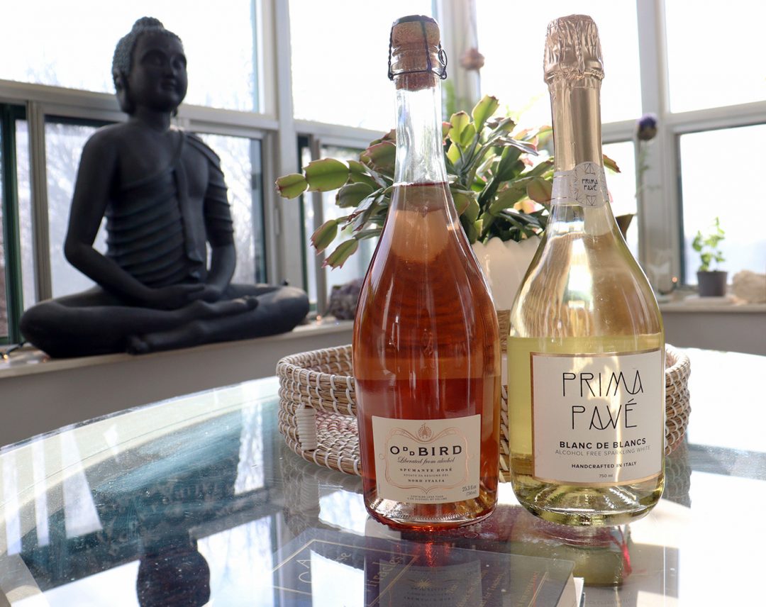 A picture of two wine bottles. one brown and one yellow, placed on a glass table with a Buddha statue in the background.