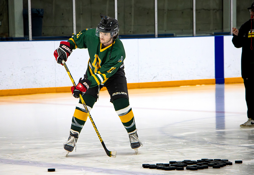 A hockey player in a green and yellow jersey winds up mid-stride and shoots a hockey puck with a yellow-and-black stick on an indoor rink. Dozens of pucks are scattered nearby as a volunteer in dark clothing stands along the sidelines observing the attempt.