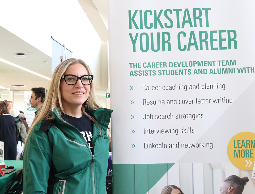 Woman with black glasses and a green jacket, poses beside a large sign that says "kickstart your career" in green text.