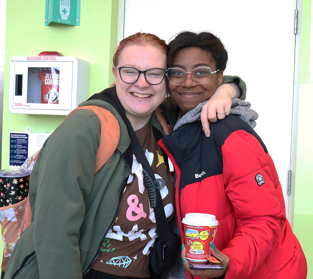 A girl in a green jacket and orange backpack hugs another girl wearing a red and black coat, holding a Tim Hortons cup.