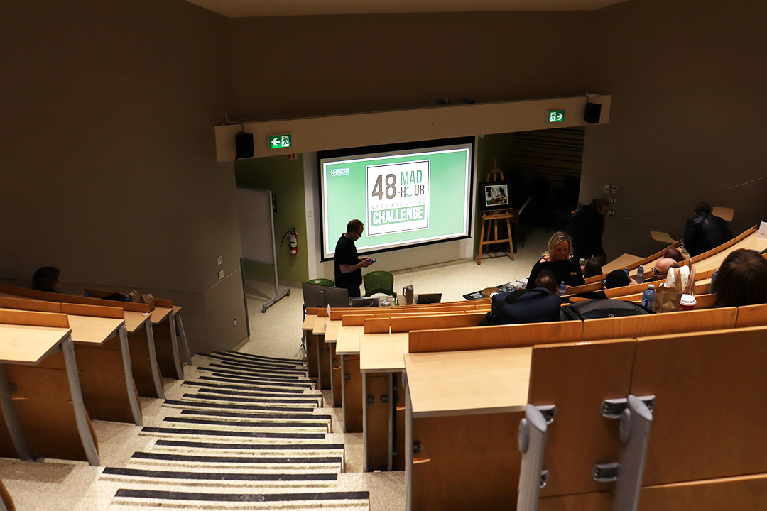 College lecture hall with stairs to the right of the image that lead down to the front of the room. Rows of wooden benches semi-full of students and staff follow the stairs downwards. A big screen sits at the button of the lecture hall with a green background, a white square sits in the middle and "MAD 48 Hour Storytelling Challenge" is written in the square.