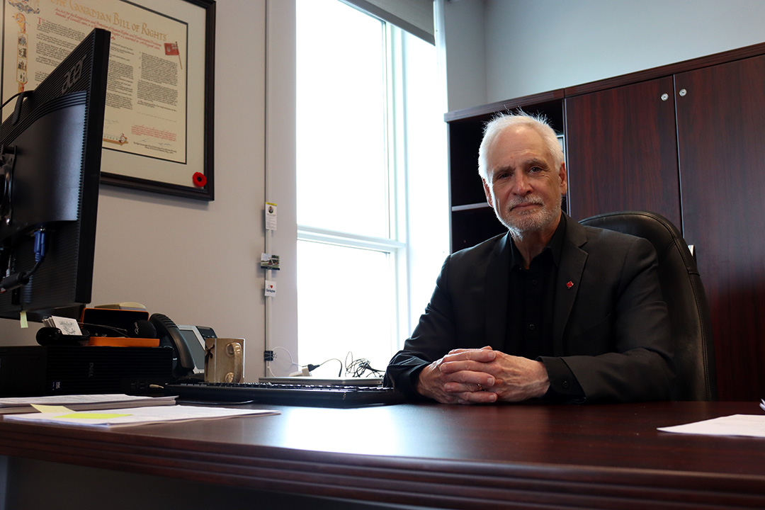 Man with short white hair in a black suit sits at his dark mahogany desk. A matching armoire with bookshelves is behind him and lights shines through a window on his left. To the far left is the back of a computer screen which is below a hanging picture of the Canadian Bill of Rights.