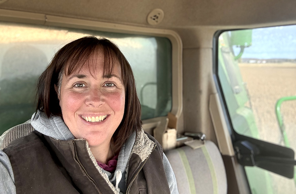 Women with dark short hair, wearing a brown and grey jacket. She's smiling at the camera and sitting in a green tractor plowing grain fields.