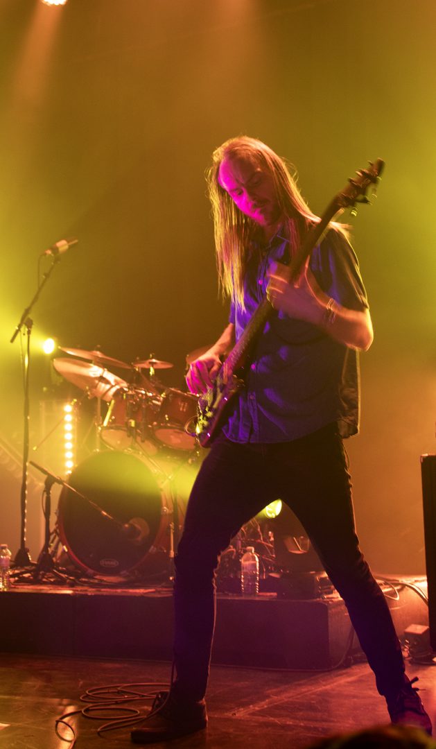 A man with long hair plays bass on stage against yellow lighting