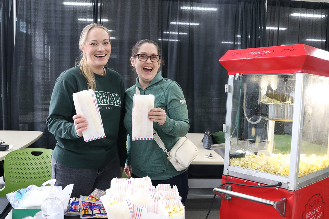 Two women wearing green shirts stand beside a red popcorn maker. They are holding bags of popcorn.