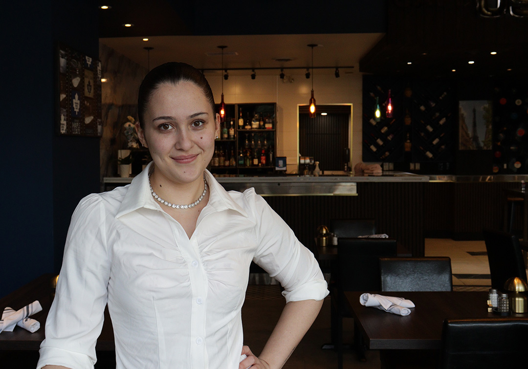 A woman wearing a white blouse and her hair tied in a slick back ponytail stands in front of tables.