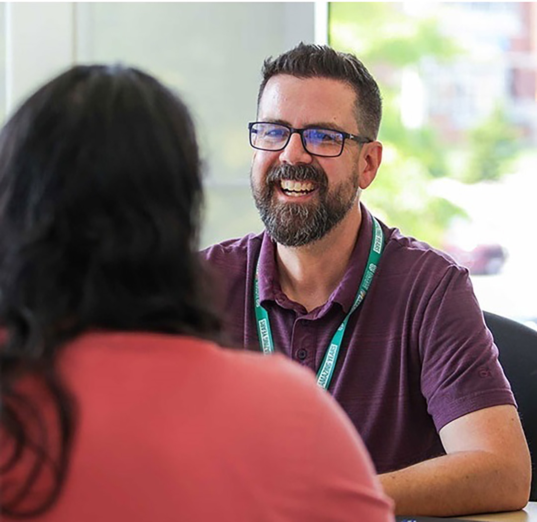 A man with glasses wearing a purple t-shirt s sitting and smiling at a woman wearing a pink shirt.