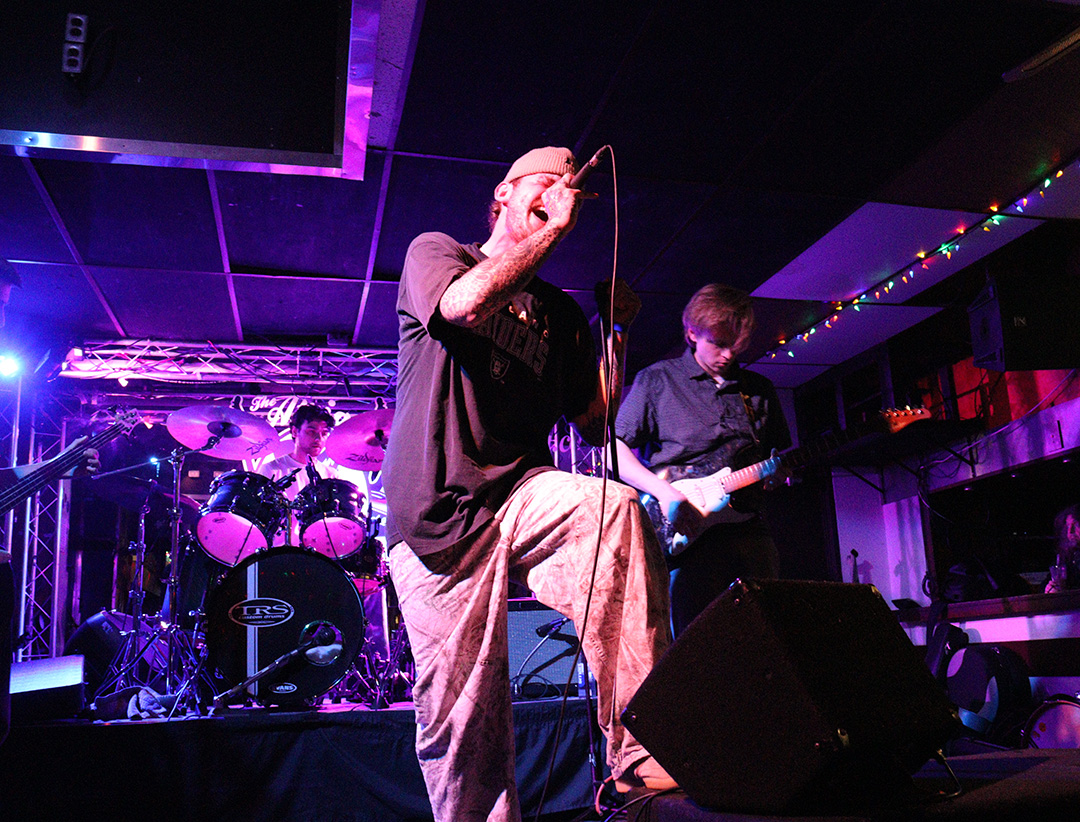 A man wearing a black T-shit and a beanie on his head sings into mic while drummer and guitarist play in the background.