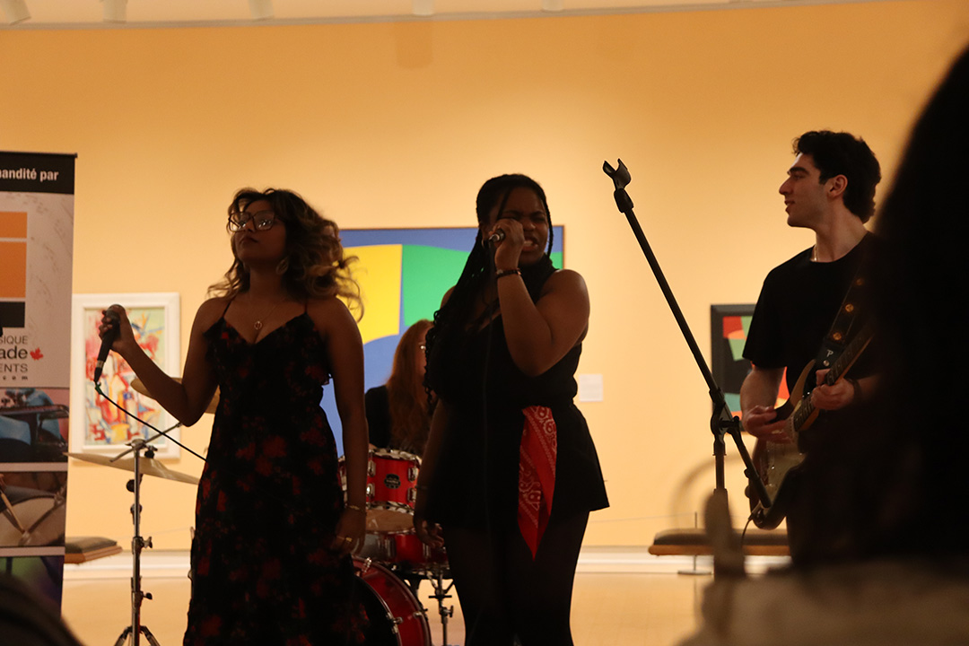 A young woman wearing a black and red flowered dress swaying while holding a mic, a second young woman wearing all black with a red bandana hanging from her skirt singing into a mic, and a young man playing gutair.