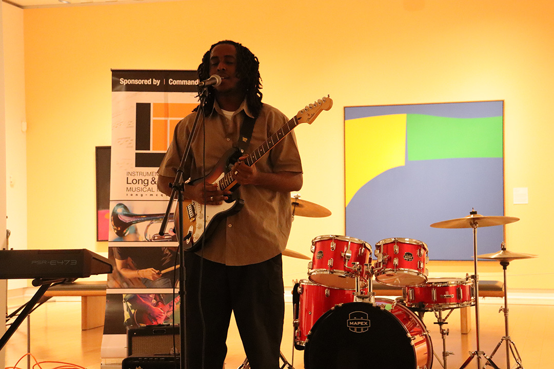 Young man with a tan button up shirt playing gutair and singing at the same time performing in an art gallery.