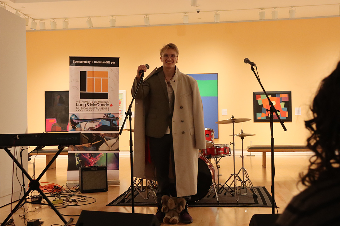 Young man poses for a photo before performing a song in an art gallery setting with an elephant plushy by his feet, while holding a microphone.