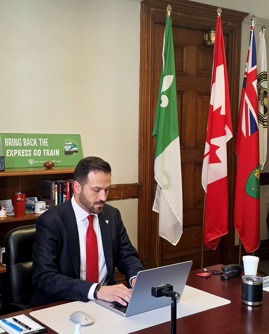A man wearing a dark blue suit, white shirt and red tie sits at a desk. There is an Express GO train banner behind him and three flags to his right.