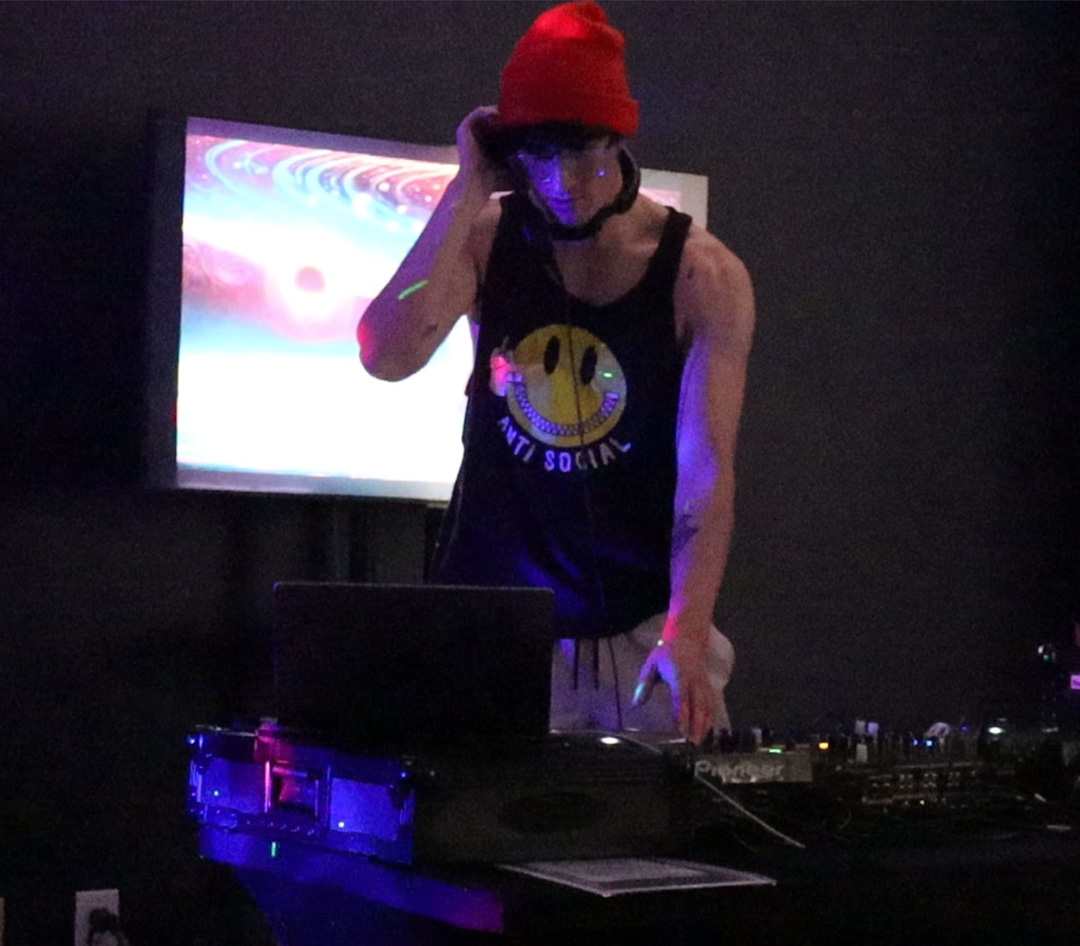 Man in an orange hat and shirt with a yellow smiley face on it uses DJ turntable in dark room.