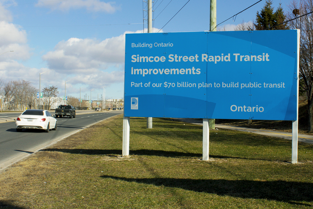 Cars pass by a sign that says "Simcoe Street Rapid Transit Improvements"