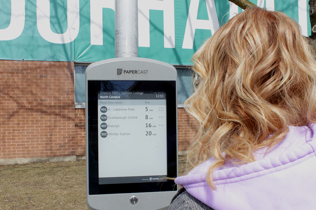 A student looks at a screen that displays the times that each bus will arrive at the stop.