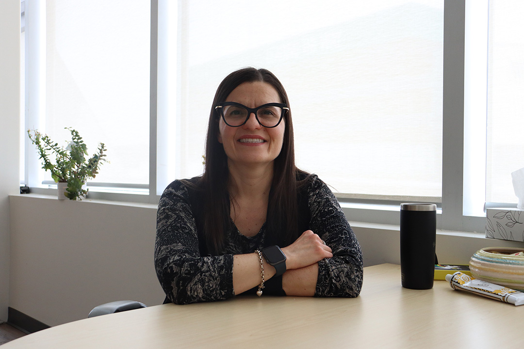 Woman wearing glasses smiles with her arms crossed while sitting at a desk infront of a bright window shining in natural light.