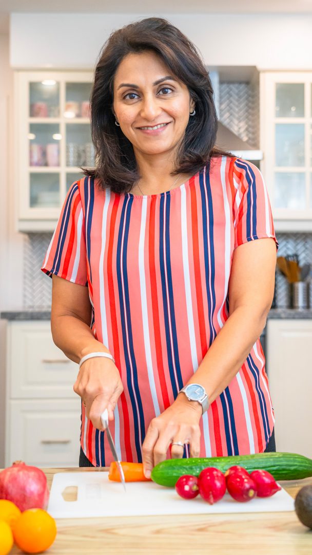A woman wearing a pink, blue, white and red striped shirt is smiling as she cuts a carrot on a cutting board.
