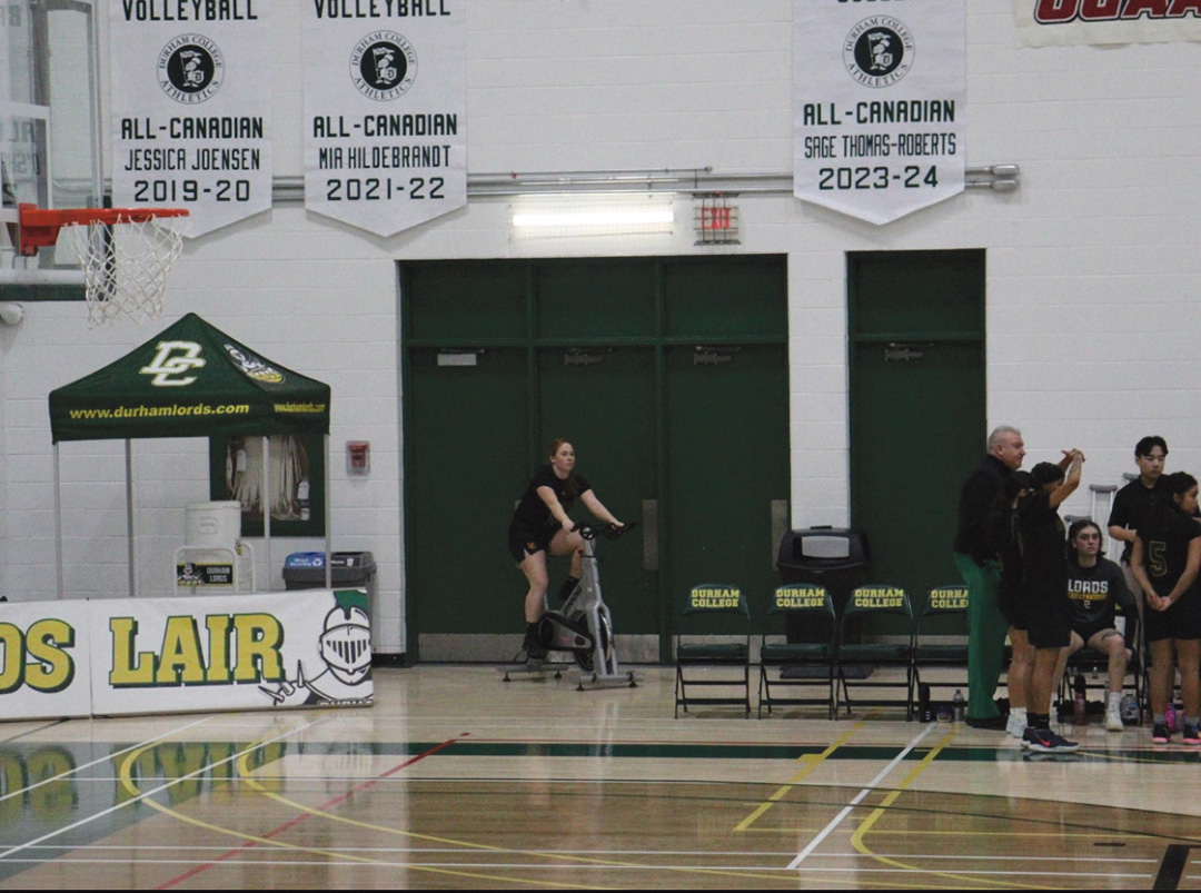 A female basketball player in a black jersey riding a bike, with other players surrounding the bench.