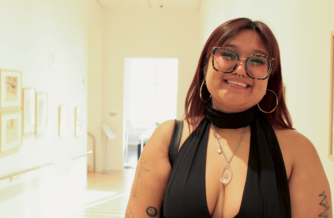 A woman in a black halter dress, hoop earrings and glasses poses for a photo in a hallway with art on the walls