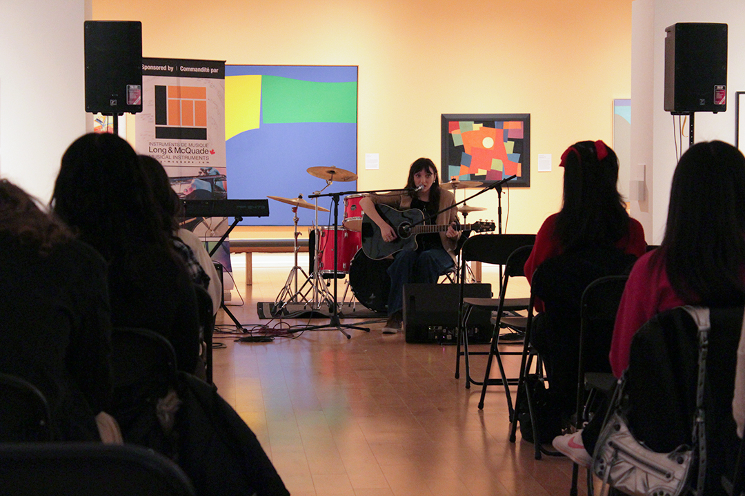An audience watches musician a young woman as she plays guitar and sings.