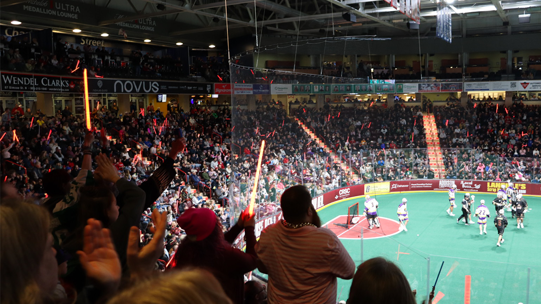 People cheer in the stands at a lacrosse game. There are players wearing black and white jerseys on the field.