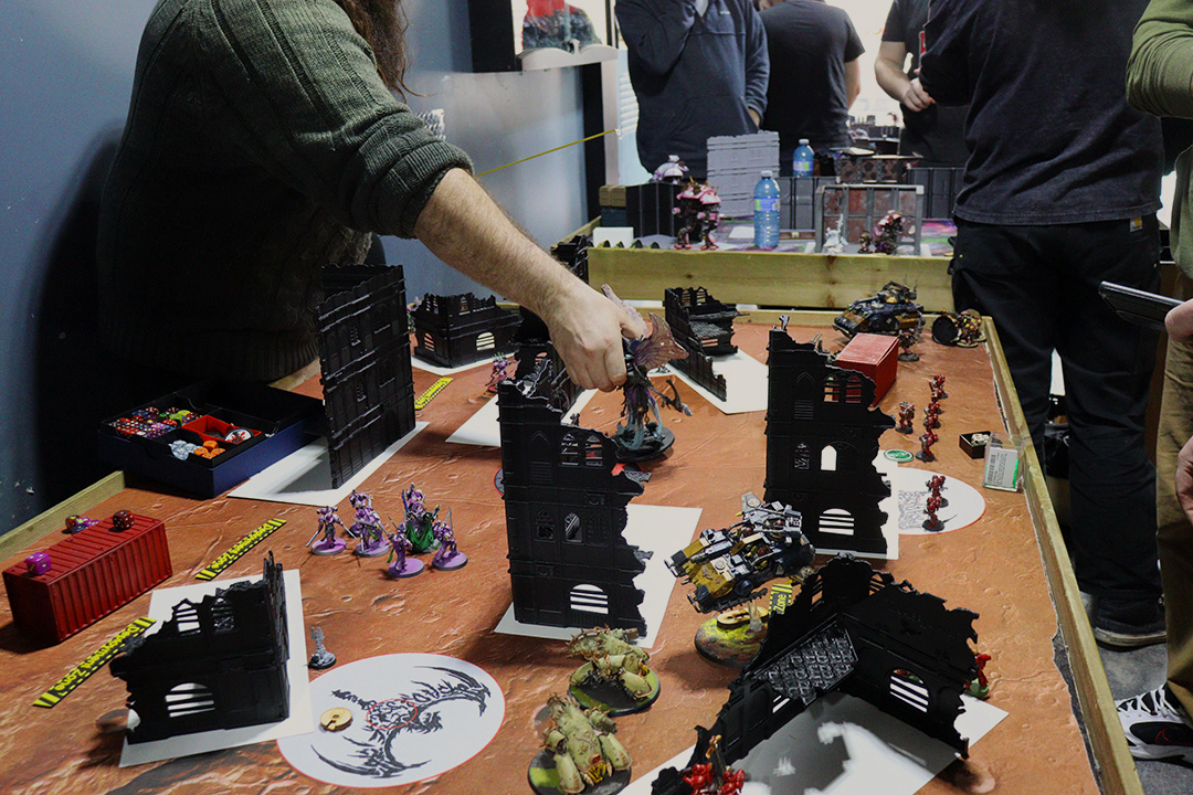 A player wearing a green knit sweater moves a Warhammer figurine on a table top. The table has an orange mat and Warhammer terrain on it. There are other players surrounding the table watching and playing.
