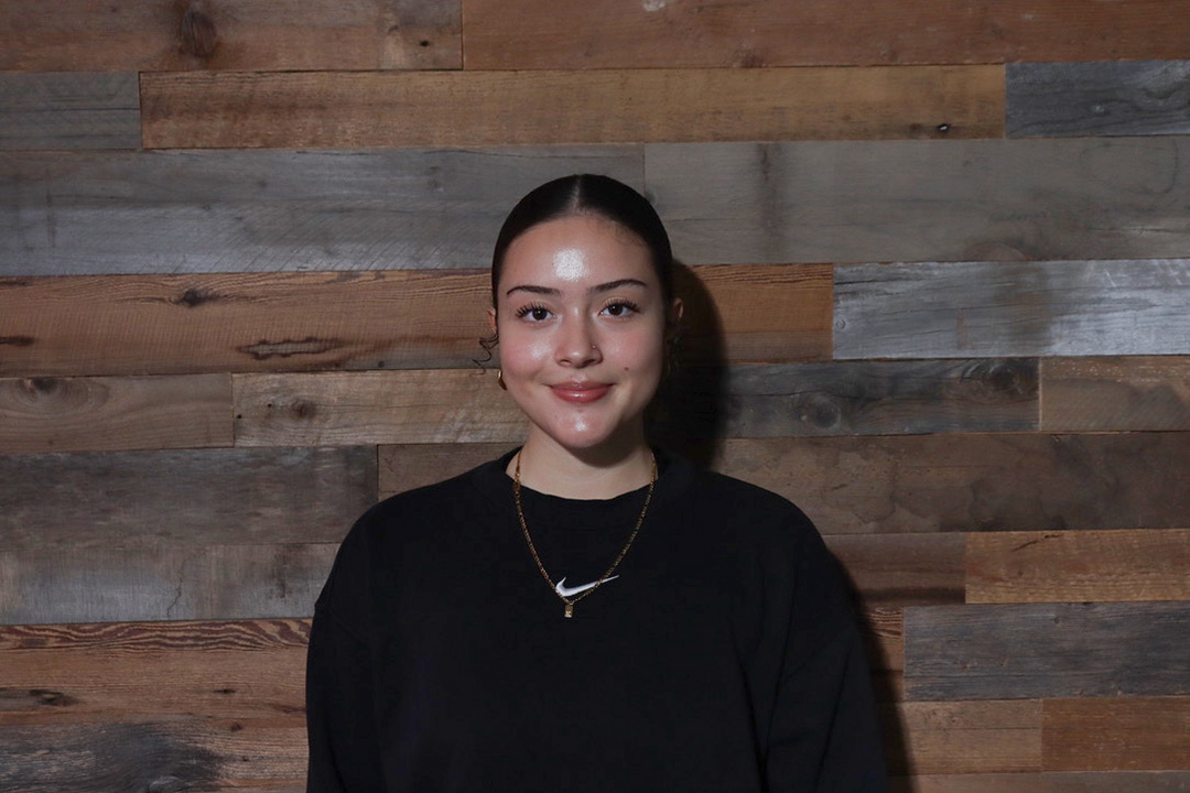 A woman in a black sweater with black hair pulled back in a ponytail stands against a barnboard wall.