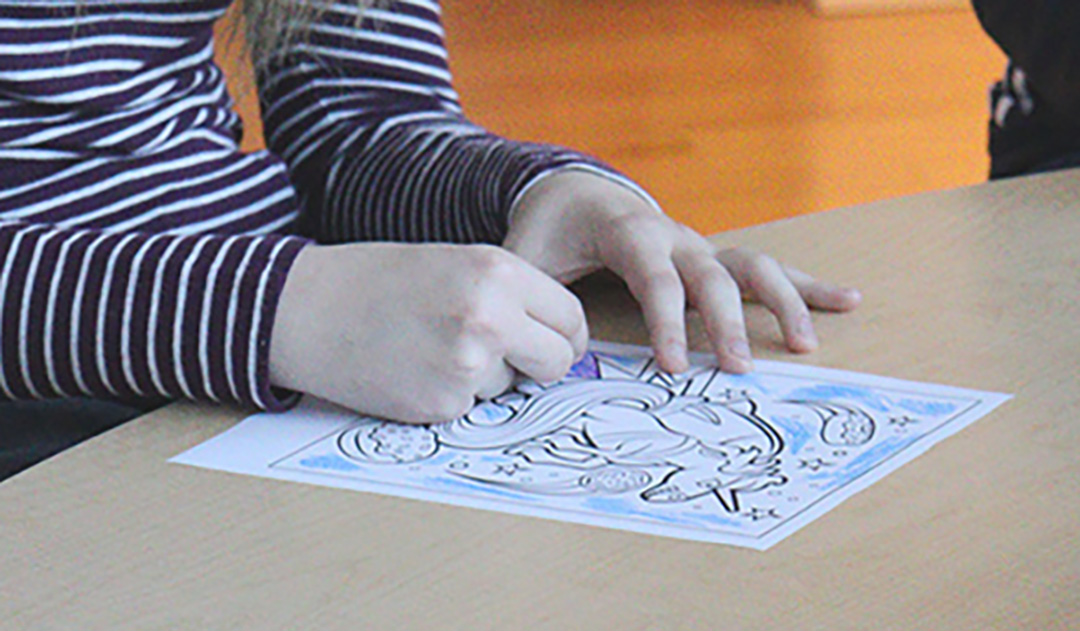 Hands on top of a colouring sheet. The person is holding a purple crayon and colouring the sheet.
