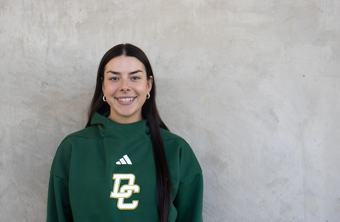 Woman with long dark brown hair wearing a dark green sports sweater with "DC" lettering smiles in front of a concrete wall.