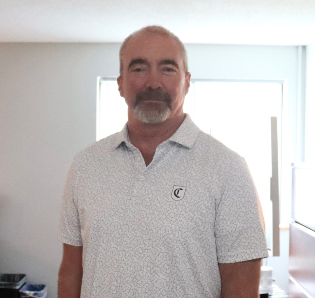 An older man stands in a white shirt in front of a white wall and open window in his office located in the Office of Campus Safety building.