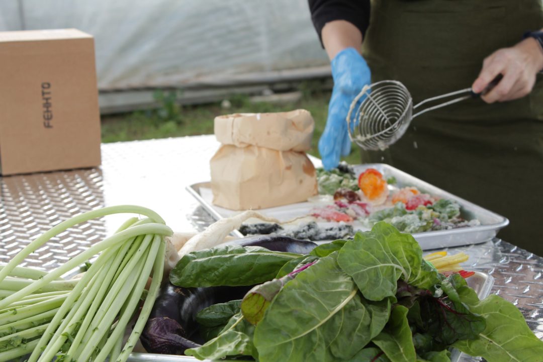 A pile of leafy vegetables sits in the foreground on top of a metal table as a person in a green apron uses a spiral egg separator to place battered vegetables on a metal tray.
